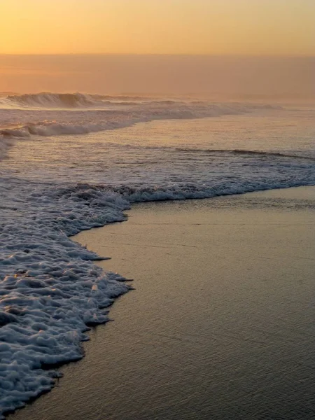A vertical shot of sea waves crashing on the sandy shore during sunset