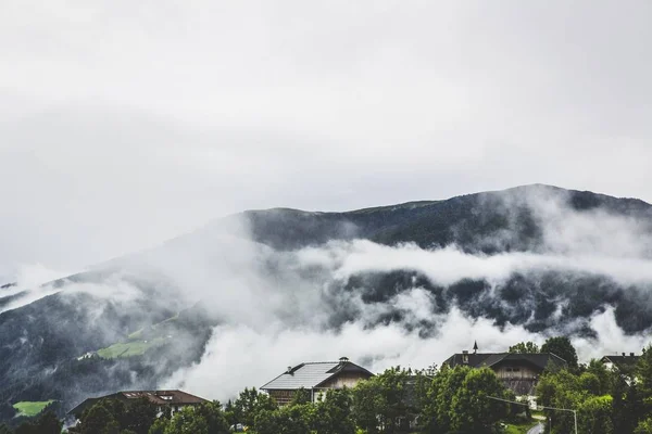 A beautiful horizontal shot of houses surrounded by trees and high mountains covered in fog at daytime