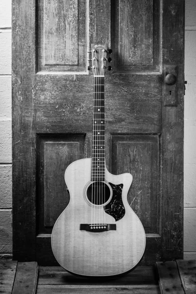 Belle prise de vue en niveaux de gris d'une guitare acoustique appuyée sur une porte en bois sur une surface en bois — Photo