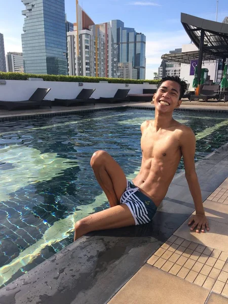 Foto vertical de un hombre feliz con traje de baño sentado junto a una piscina al aire libre cerca de edificios — Foto de Stock