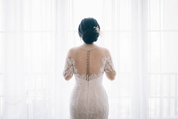 白いカーテンの近くにレースのウェディングドレスを持つ美しい花嫁の水平ショット — ストック写真
