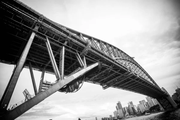 Low angle greyscale shot of a bridge surrounded by high building near the sea under beautiful sky — Stock Photo, Image