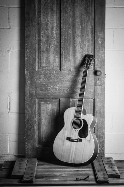 Красивая фотография акустической гитары в оттенках серого, прислоненная к деревянной двери на деревянной поверхности — стоковое фото