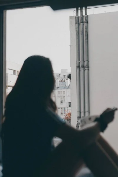 Силуэт женщины, сидящей у окна и смотрящей на здания — стоковое фото