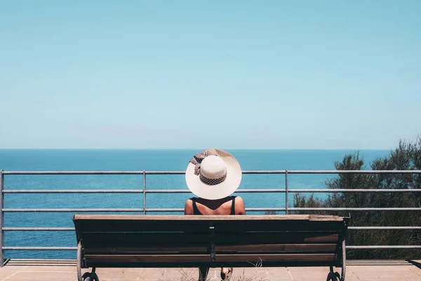 Vrouw met een witte strohoed op zittend op een bankje kijkend naar de zee onder de blauwe lucht — Stockfoto