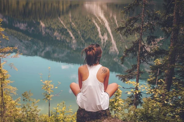 一位年轻女子坐在湖边岩石上 倒映着青山的高角镜头 — 图库照片