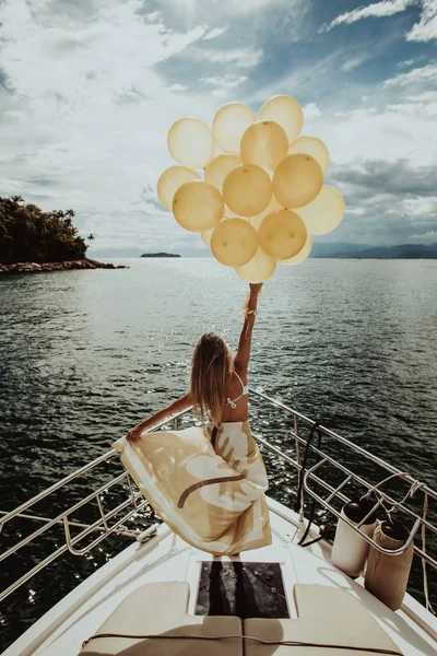 Tiro vertical de una mujer en un vestido de pie en un yate, sosteniendo globos de oro mientras navega — Foto de Stock
