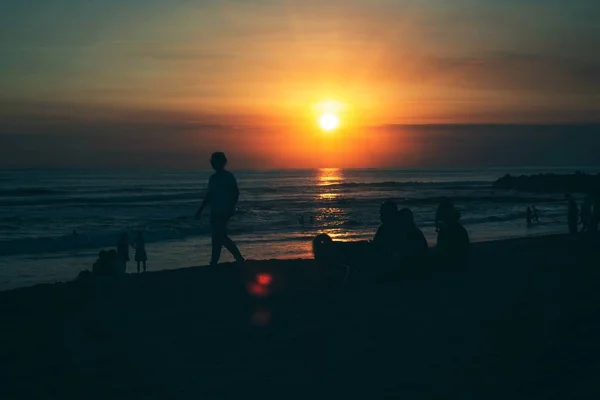 Silhueta de pessoas na praia desfrutando da bela vista do pôr do sol perto do mar à noite — Fotografia de Stock