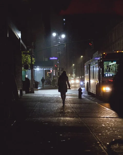 Вертикальный снимок человека, идущего по тротуару возле автобуса и зданий в ночное время — стоковое фото