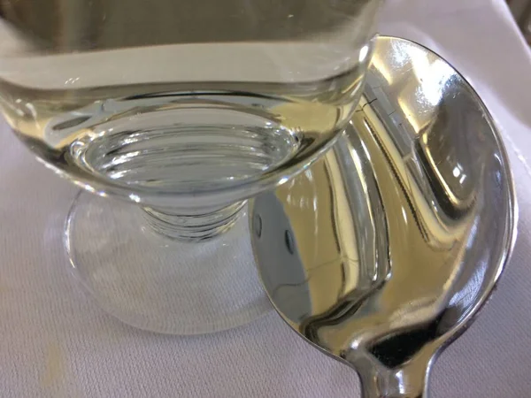 Closeup shot of a clear glass of water and a silver teaspoon next to it on a white tablecloth — ストック写真