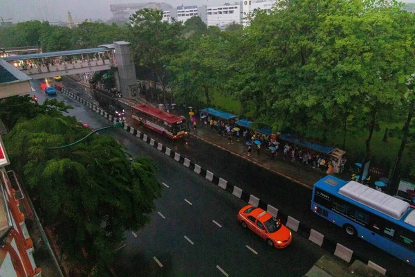 Véhicules dans la rue entourés d'arbres et de personnes se tenant sous l'abri d'arrêt de bus pendant la pluie — Photo