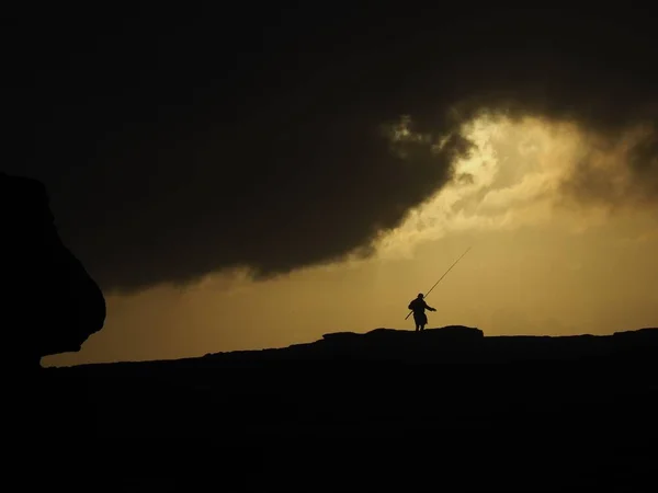 Silhouette d'une personne avec une canne à pêche debout sur la colline sous un ciel nuageux jaune — Photo