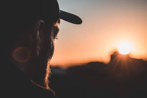 Foto superficial focada no perfil de um homem usando um boné e olhando para o pôr-do-sol borrado — Fotografia de Stock