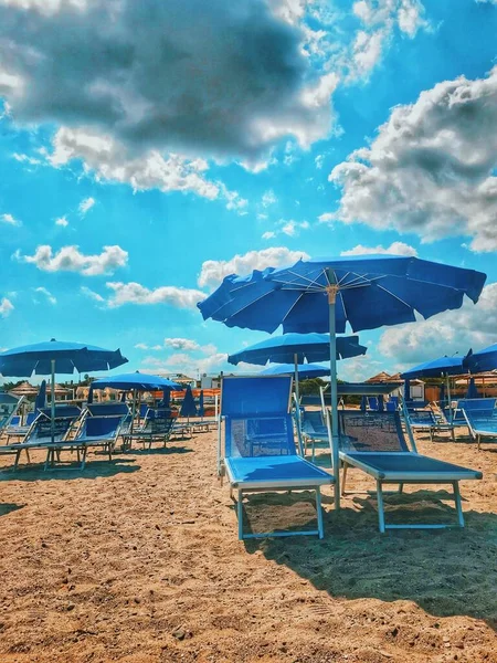 Вертикальний знімок синіх парасольок і шезлонгів на пляжі під захоплюючим хмарним небом — стокове фото