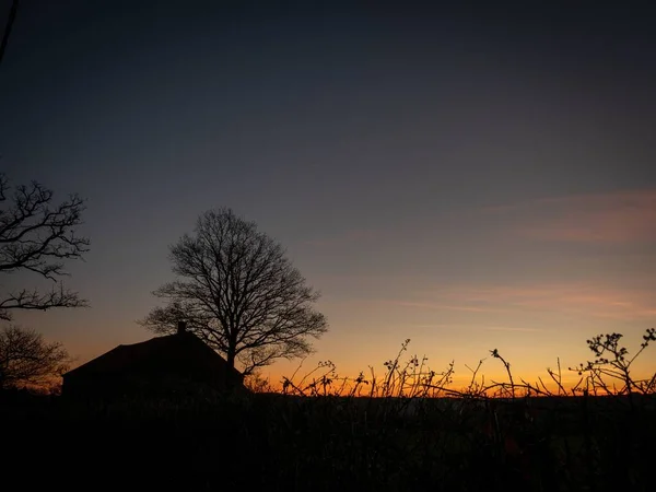 Silueta de una casa cerca de árboles sin hojas con un hermoso cielo en el fondo al atardecer — Foto de Stock