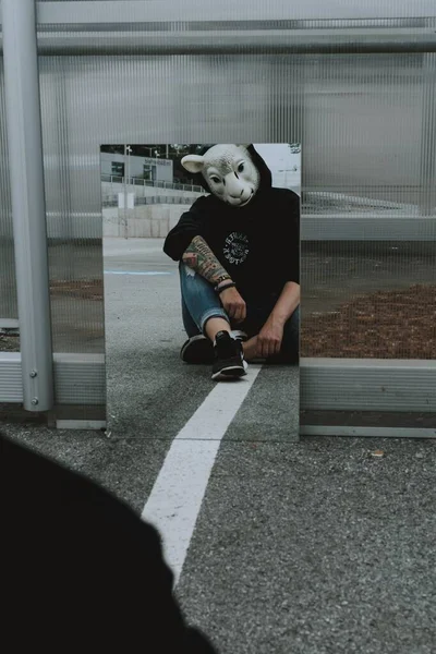 Black hooded person with a sheep mask staring at his reflection in the mirror on a pavement — Stock Photo, Image