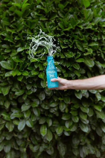 En person som strör vatten i en blå flaska med ett plastfritt tryck framför gröna lagerhus — Stockfoto