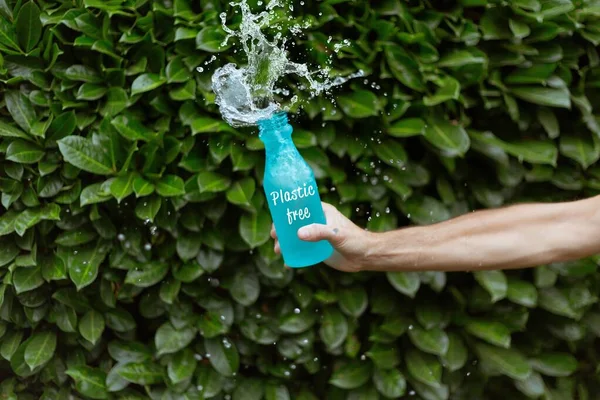 En person som strör vatten i en blå flaska med ett plastfritt tryck framför gröna lagerhus — Stockfoto