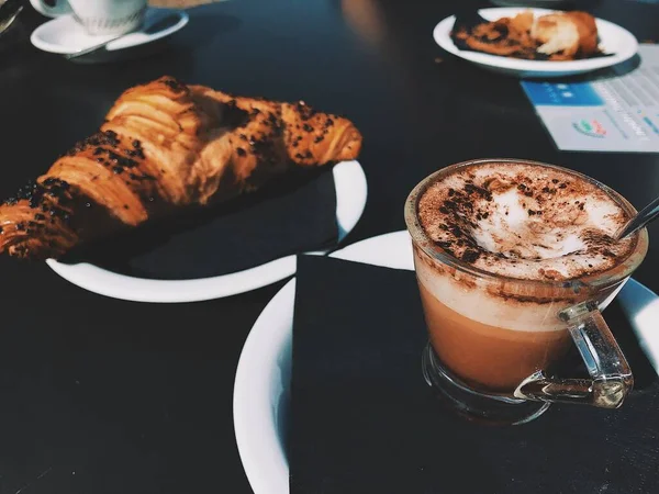 Closeup shot of a clear glass cup filled with cappuccino beside a croissant — Stock Photo, Image