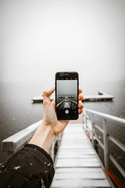 Fotografía vertical de una persona con un smartphone tomando una foto de un muelle cerca del hermoso mar —  Fotos de Stock