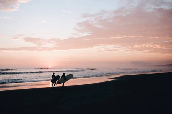 夕暮れ時に二人のサーファーのシルエットが海岸にサーフボードを運んでいる — ストック写真