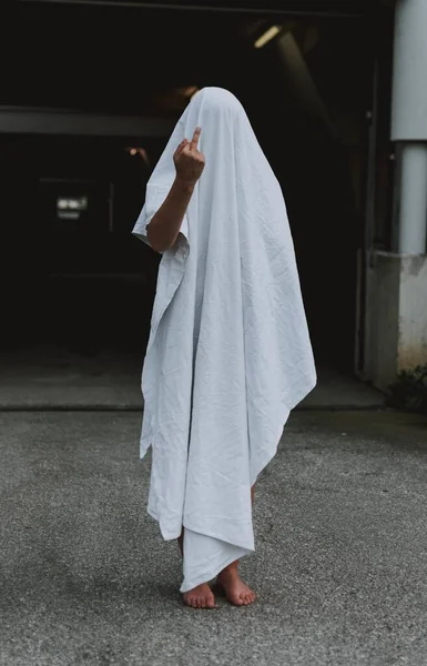 Tiro vertical de uma pessoa descalça usando lençol fantasma branco mostrando o dedo médio — Fotografia de Stock