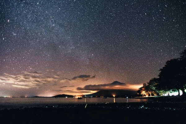 A wide shot of the beautiful sea surrounded by green trees and mountains under the breathtaking starry night sky