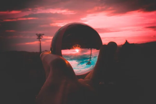 Una persona sosteniendo una bola de vidrio con el reflejo del cielo colorido y la hermosa puesta de sol — Foto de Stock