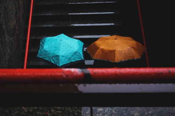 Horizontal shot of two persons going down the stairs while holding a cyan and brown umbrellas