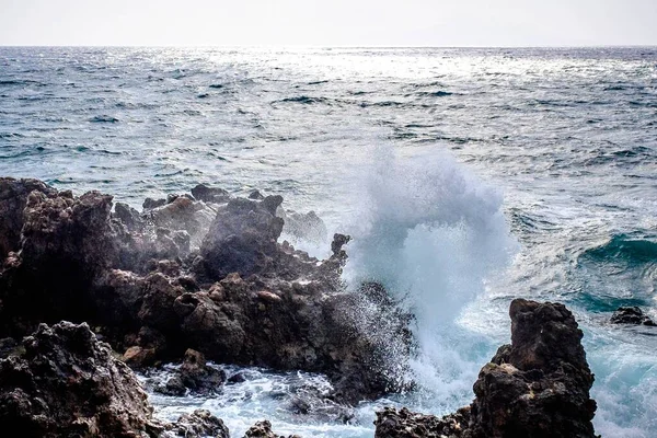 A horizontal shot of a rocky shore near a body of water with waves splashing the rocks during the day