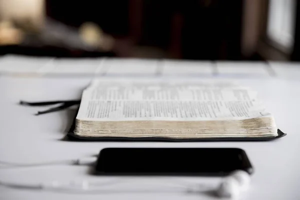 在背景模糊的智能手机附近拍摄的一本打开的圣经的特写镜头 — 图库照片