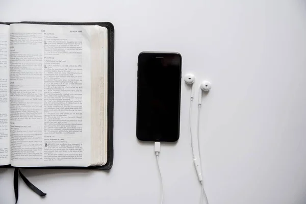 Overhead-Aufnahme eines Smartphones mit Kopfhörer in der Nähe einer offenen Bibel auf einer weißen Oberfläche — Stockfoto