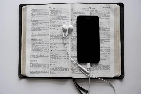 Overhead shot of a smartphone and headphones on an opened bible on a white surface — Stock Photo, Image