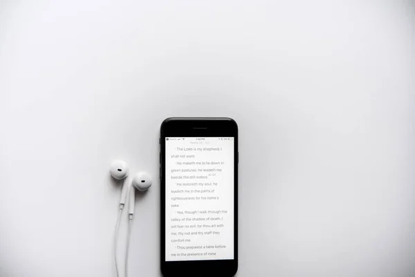 Верхний снимок библии на смартфоне рядом с наушниками на белой поверхности — стоковое фото