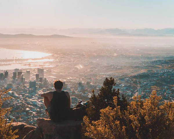 Homem sentado no topo da rocha admirando a vista da cidade abaixo — Fotografia de Stock