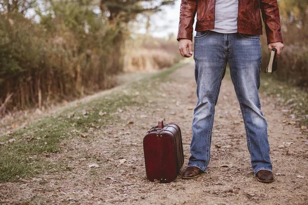 Eski bavulunun yanında boş bir yolda duran ve geçmişi bulanık bir şekilde İncil 'i tutan bir erkek. — Stok fotoğraf