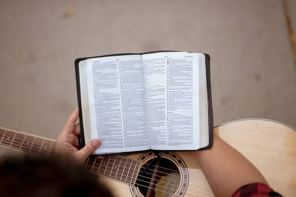 Накладний знімок людини, що сидить зі своєю гітарою читає Біблію з розмитим тлом — стокове фото