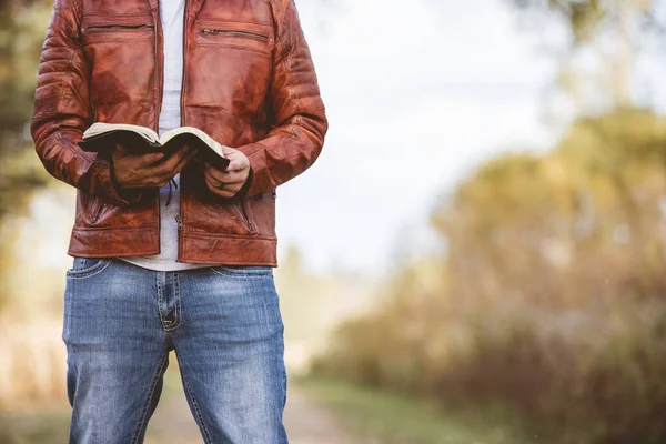 Hombre usando una chaqueta de cuero de pie en un camino vacío y leyendo la Biblia con un fondo borroso — Foto de Stock