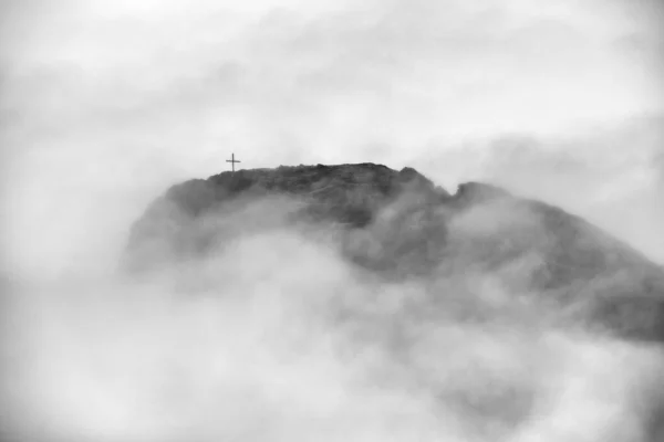 Hermosa foto de una montaña con una cruz en la parte superior rodeada de niebla — Foto de Stock