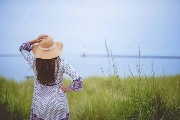 Žena nosí letní klobouk s rukou na boku, zatímco stojí v travnatém poli střílel zezadu — Stock fotografie