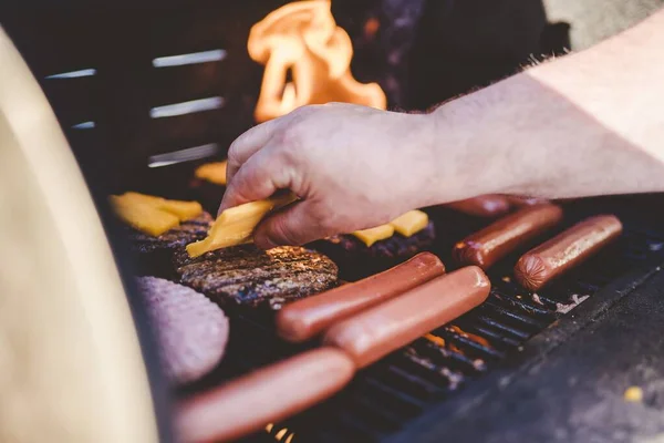 Zbliżenie osoby umieszczającej ser na hamburgerze wołowym na grillu w pobliżu hot dogów — Zdjęcie stockowe
