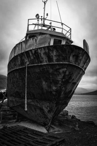 Tiro vertical de um velho navio enferrujado na costa perto da água sob céu nublado em preto e branco — Fotografia de Stock