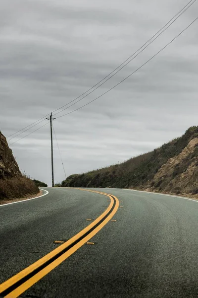 Tiro vertical de uma estrada de dois lados cercada por colinas com o céu cinza nublado no fundo — Fotografia de Stock