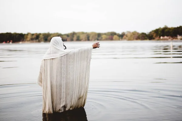 Persoon die een bijbels gewaad draagt en in het water staat met een hand omhoog geschoten van achteren — Stockfoto