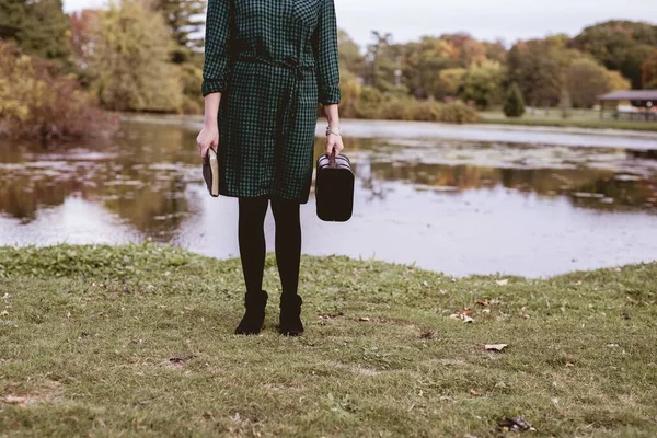 Female standing on a grassy field near water while holding her bible and an old suitcase — Stock Photo, Image