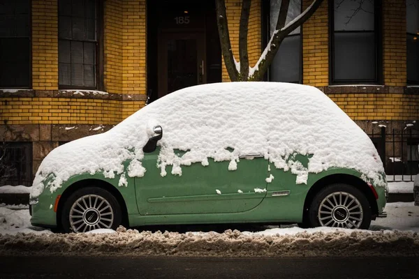 Green car half-covered with snow parked in front of a building with yellow walls in the background — Stock Photo, Image