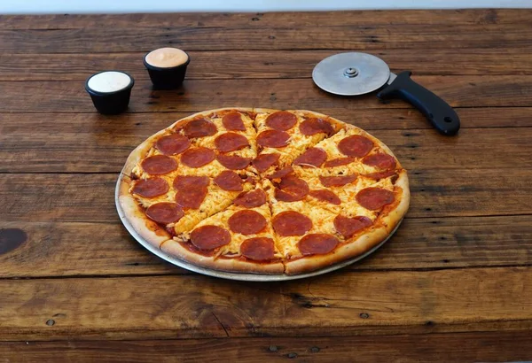 A closeup shot of a pepperoni pizza with sauces and a cutter on a wooden table