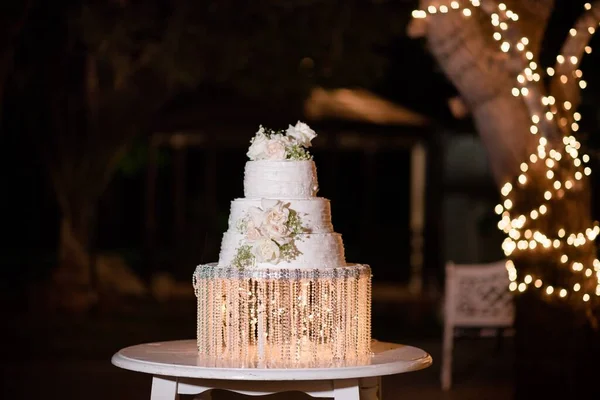 A three tiered wedding cake displayed at an outdoor reception at  night, adorned with fresh roses on a cake stand with crystals