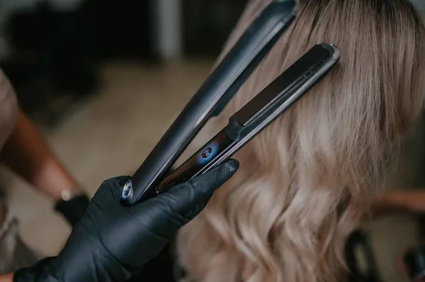 A black hair straightener held by a person with black gloves and blonde wavy hair