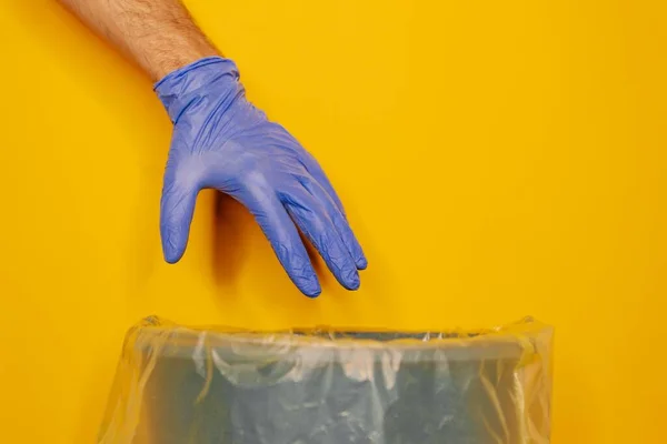 在黄色背景下 一个男人在垃圾桶上方手拿着医用手套的特写镜头 — 图库照片